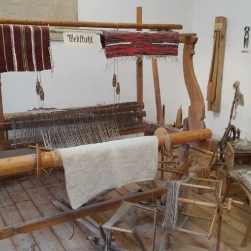 Ein alter Handwebstuhl aus Holz. Rundherum und an der Wand hängt altes Zubehör das man für das Weben benutzt hat. Der Webstuhl steht in einem Museum und ist nicht mehr in Betrieb.