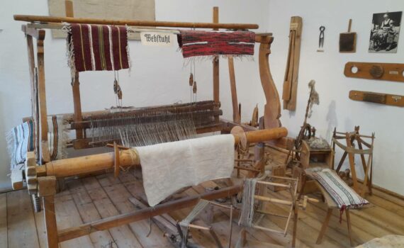 Ein alter Handwebstuhl aus Holz. Rundherum und an der Wand hängt altes Zubehör das man für das Weben benutzt hat. Der Webstuhl steht in einem Museum und ist nicht mehr in Betrieb.